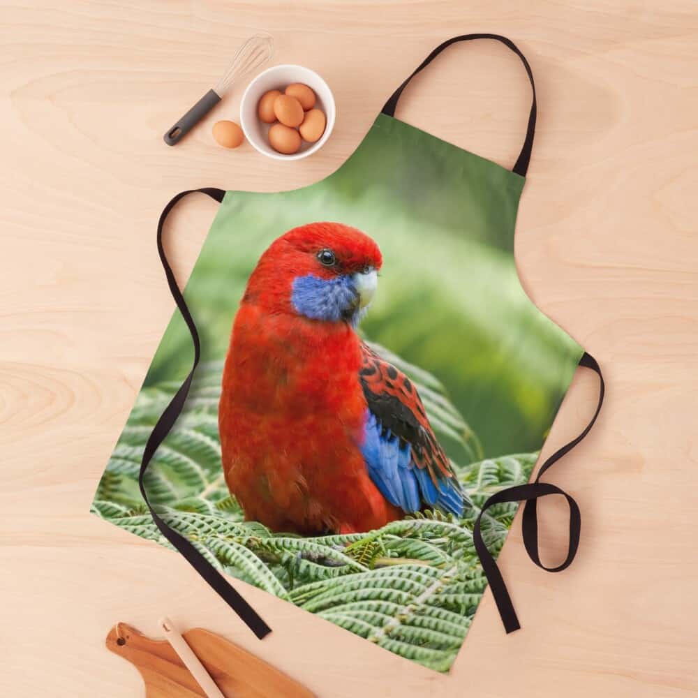 an apron with an image of a Crimson Rosella on it layed out on a table with a bowl of eggs, a whisk, and a chopping board