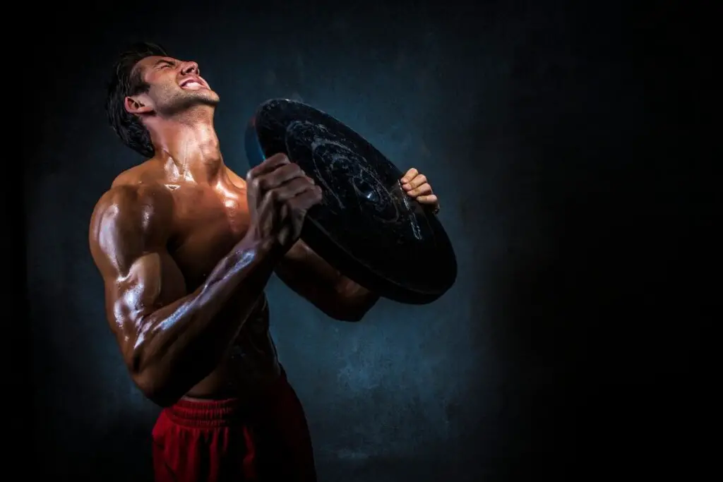 a shirtless man in red pants struggling to lift a heavy circular weight.