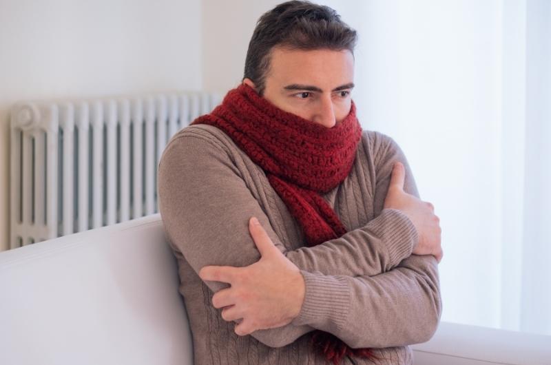 a man sitting in a bright room wearing a jumper and a scarf and shivering