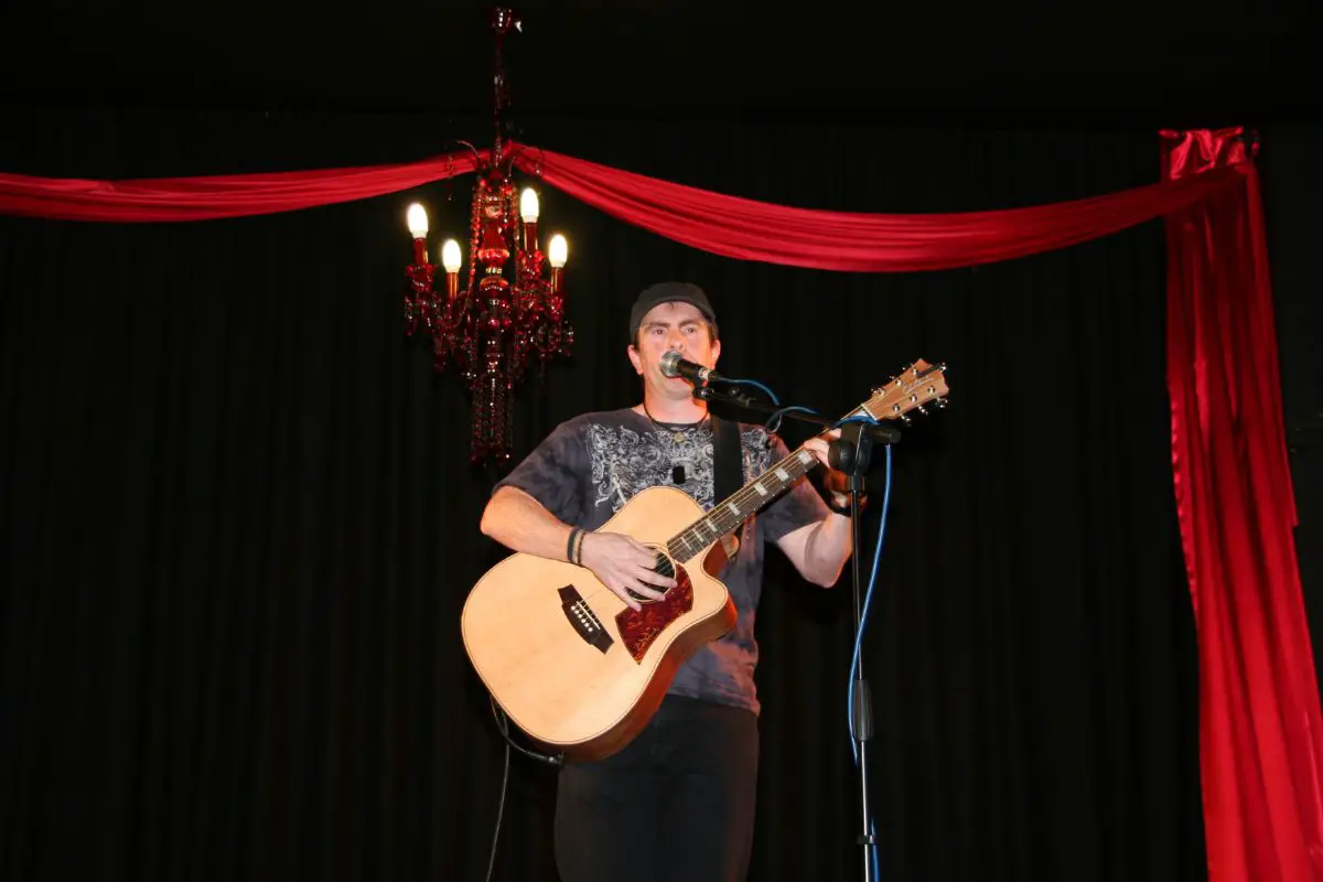 a male guitarist on stage with a chandelier and bright red curtain hanging behind him